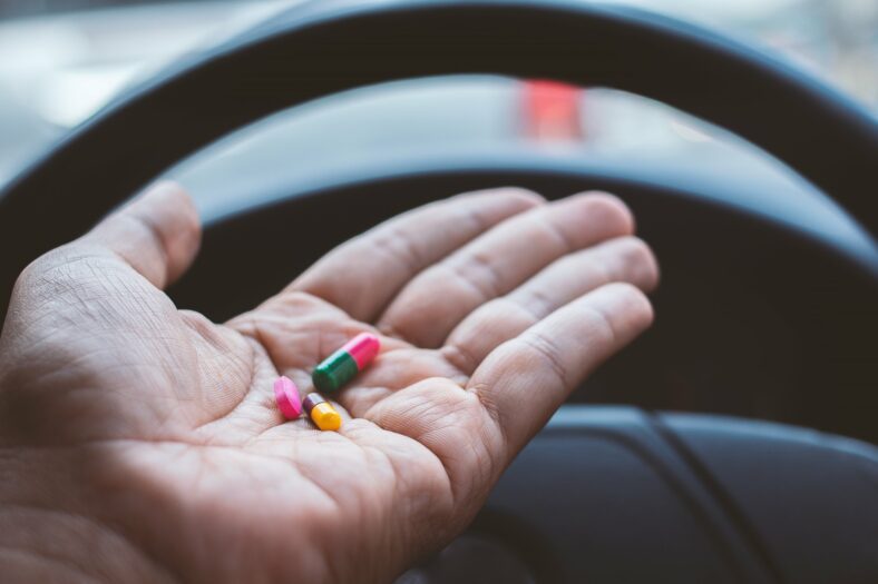 Arzneimittel_im_Strassenverkehr.jpg Eine Hand hält bunte Kapseln über dem Lenkrad eines Fahrzeugs.