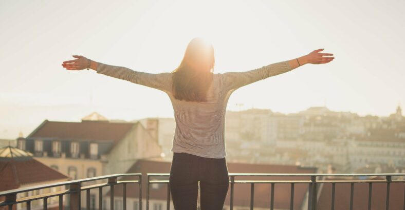 Gesundes_Immunsystem.jpg Eine Person mit ausgestreckten Armen steht auf einem Balkon und blickt in die Sonne über einer Stadt.