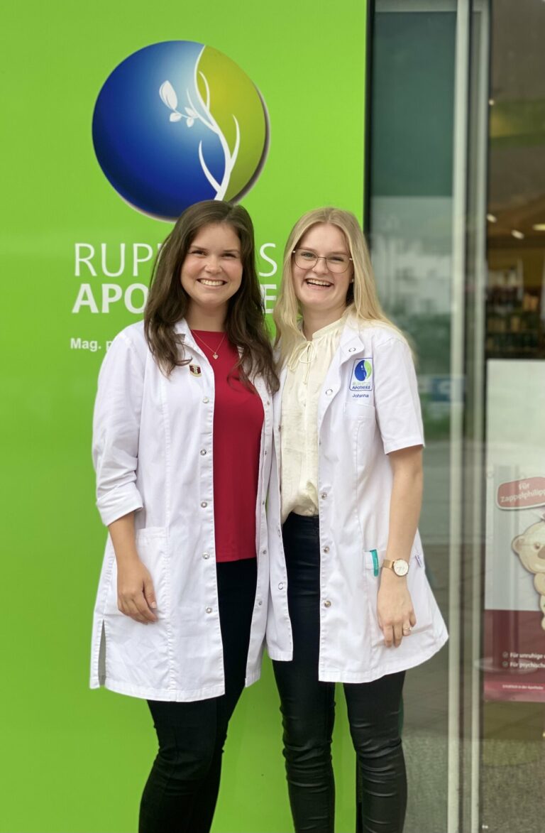 Zwei Apothekerinnen in weißen Kitteln stehen vor einer grünen Wand mit dem Logo der Apotheke.
