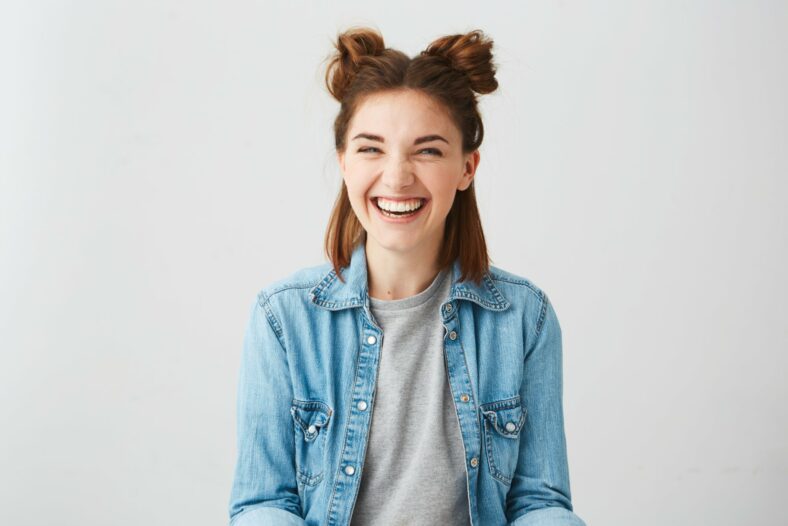 Lachen.jpg Lachende junge Frau mit Duttfrisur, trägt eine Jeansjacke über einem grauen T-Shirt, vor einem hellen Hintergrund.