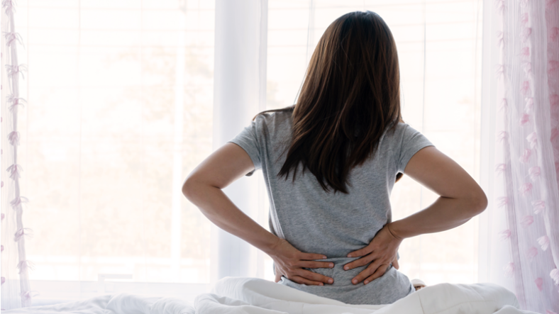 Ruecken_web.png Eine Person mit langen Haaren sitzt auf einem Bett und hält sich den Rücken vor einem Fenster.