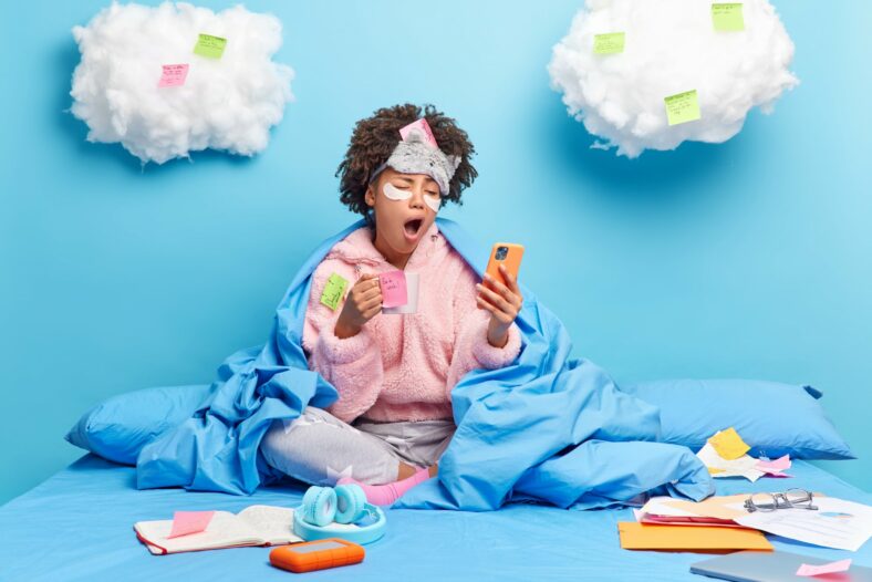 Soforttipps_gegen_Muedigkeit.jpg Eine Person in einem blauen Bett, umhüllt von einer Decke, schaut müde auf ihr Handy und hält eine Tasse in der Hand. Wolken mit Haftnotizen im Hintergrund.