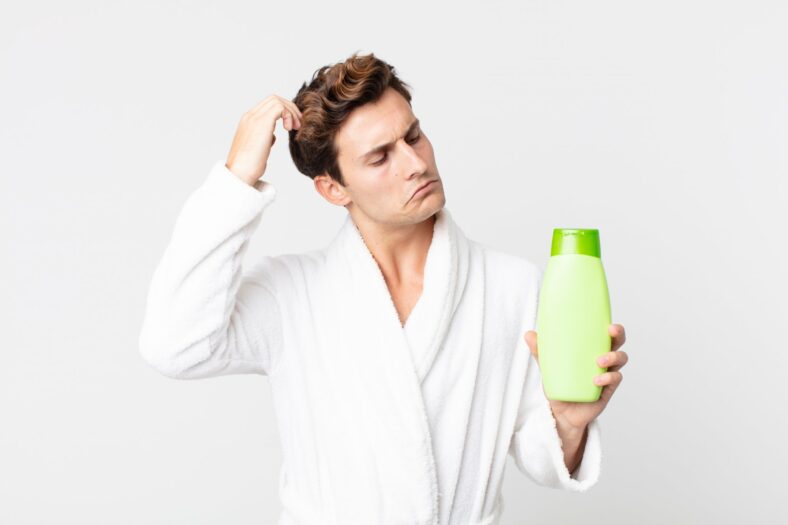 Trockene_Kopfhaut_Pflege.jpg Ein Mann in einem weißen Bademantel hält eine grüne Shampoo-Flasche und sieht nachdenklich aus.