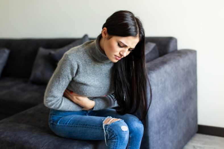 endometriose.jpg Eine Frau mit langen, dunklen Haaren sitzt auf einem grauen Sofa und hält sich den Bauch, offensichtlich unwohl.