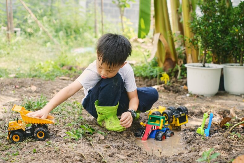 wie-viel-dreck-ist-gesund.jpg Ein Junge im Garten spielt mit verschiedenen Spielzeugfahrzeugen, darunter ein Lkw und ein Bagger, während er in matschigem Boden sitzt.